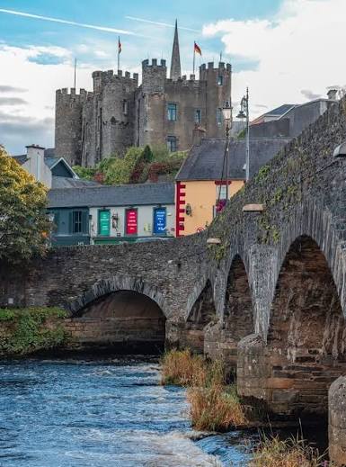 A stone bridge spans a river, with colorful buildings and a castle in the background under a partly cloudy sky